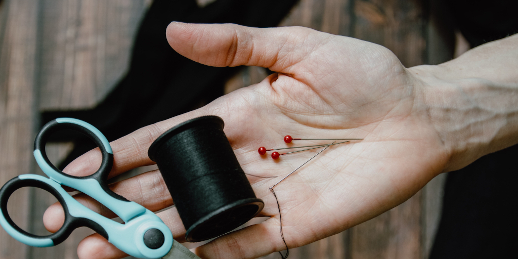 A Hole in November. Photo by Kelly Sikkema on Unsplash. Shows an open hand holding sewing pins, a reel of dark thread and scissors.