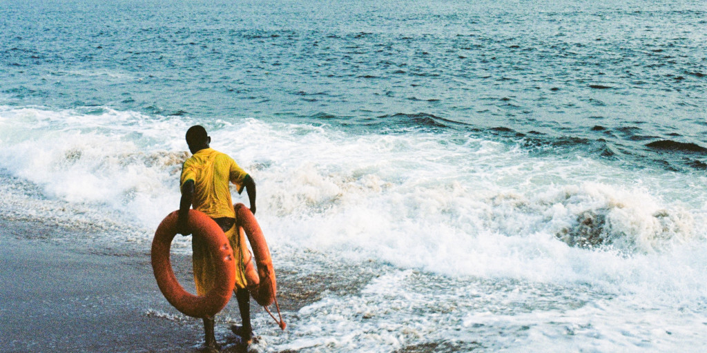 Journey. Photo by Aude-Andre Saturnio on Unsplash. Shows a man carrying two orange lifebelts by the sea.